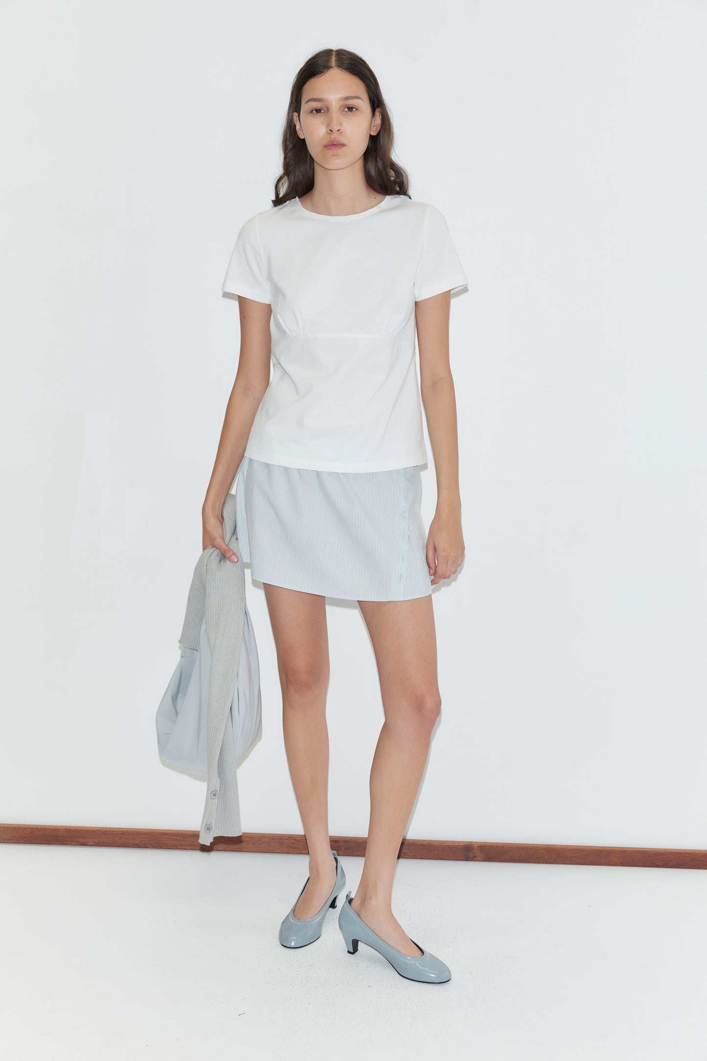 A woman stands against a plain white background wearing the Tape Tee - White by Deiji Studios, a mist stripe skirt, and blue heels, holding a light blue jacket and bag in her left hand.