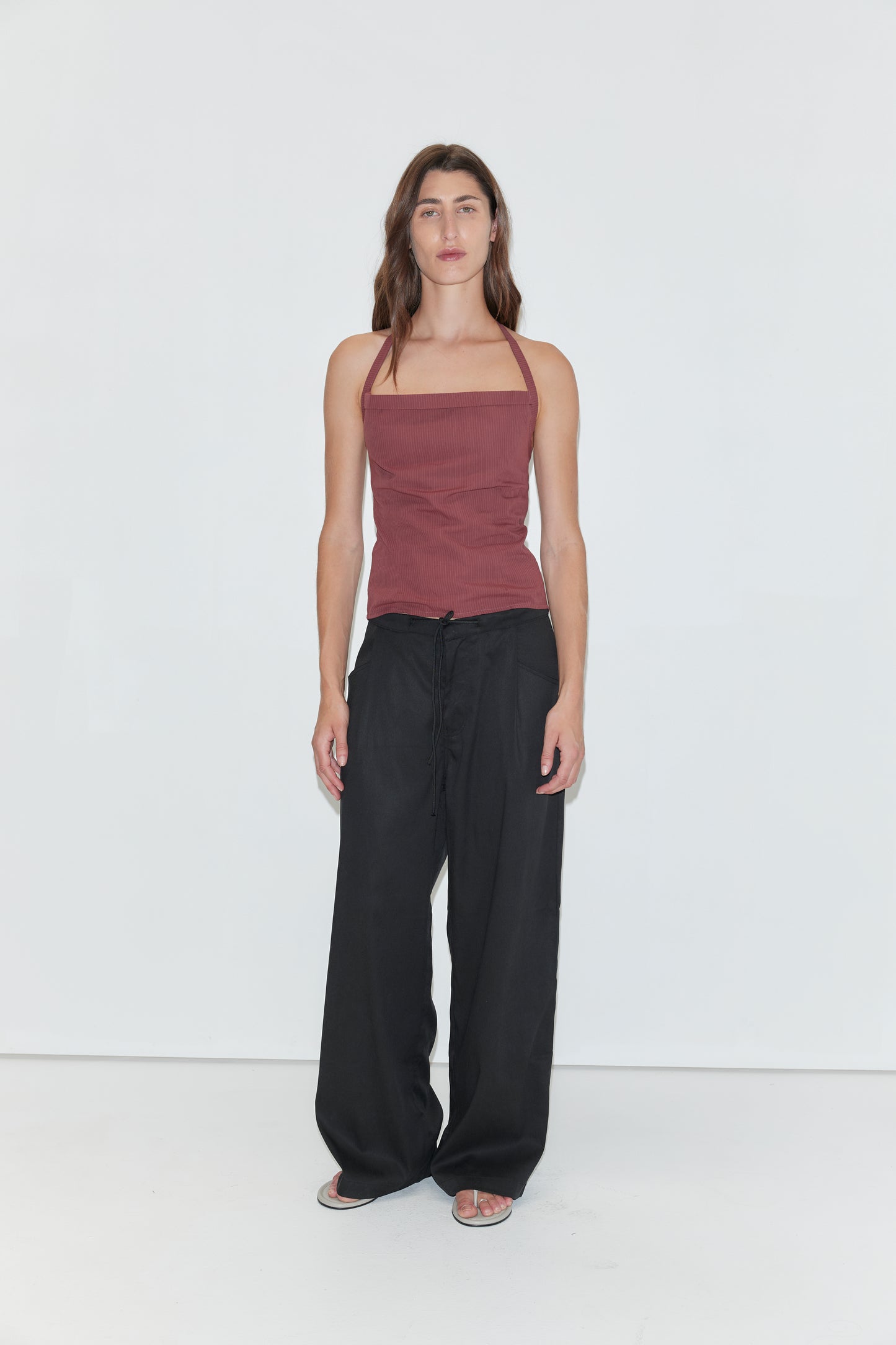 A person with long brown hair stands against a plain white background, wearing the Folded Top in Rust Stripe by Deiji Studios, paired with loose black pants and sandals.