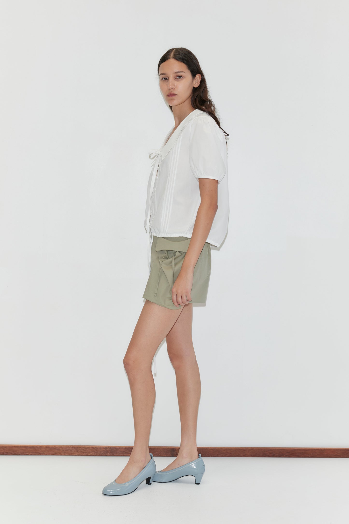 A woman with long brown hair and a neutral expression stands sideways against a white background, wearing a white short-sleeved top with ties, Micro Pocket Short - Stone Green by Deiji Studios, and light blue low-heeled shoes.
