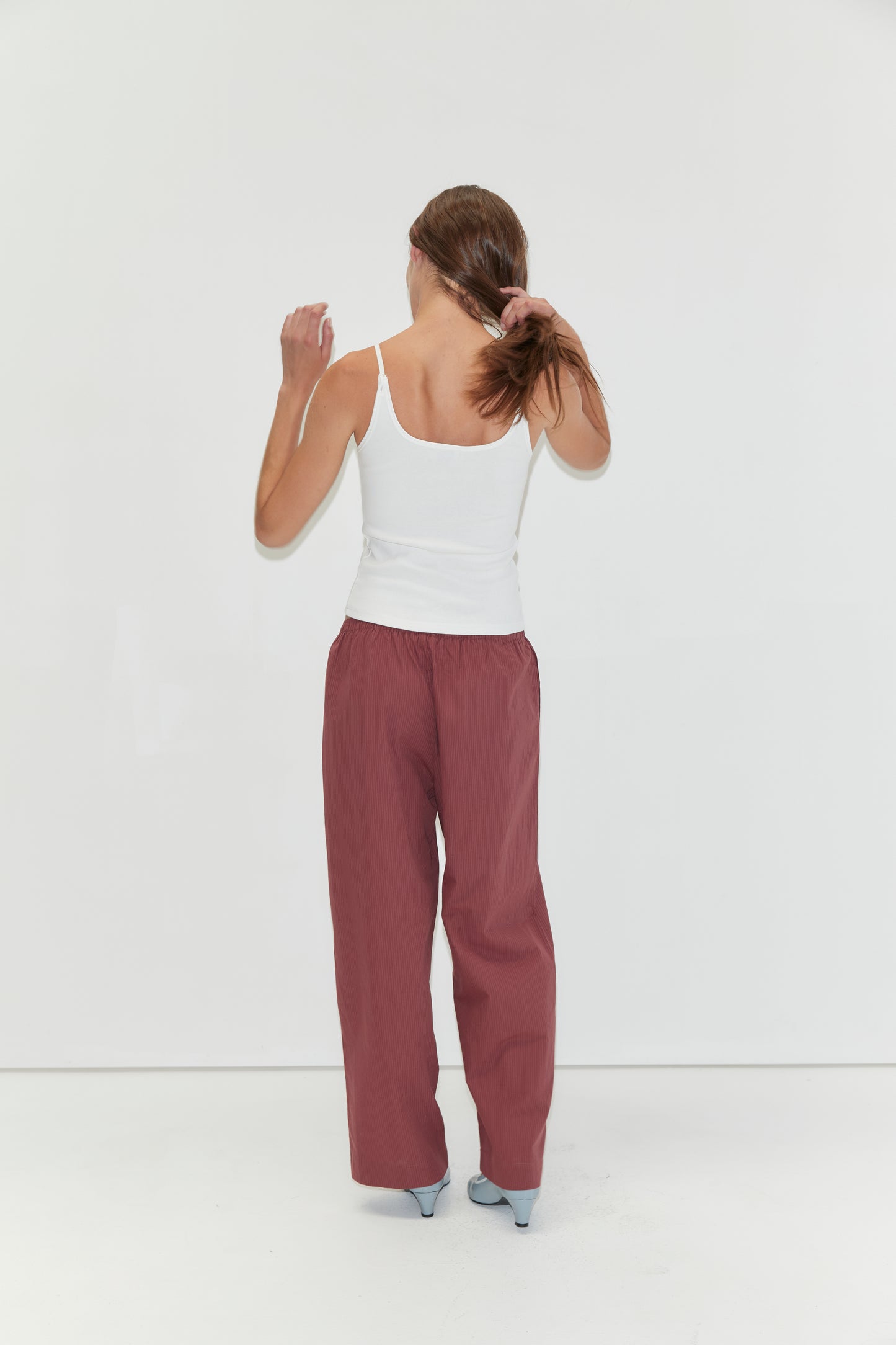 A woman stands facing a white wall, holding her brown hair in a ponytail. She wears the Rib Jersey Tank in White from Deiji Studios, loose maroon pants, and light blue heels.