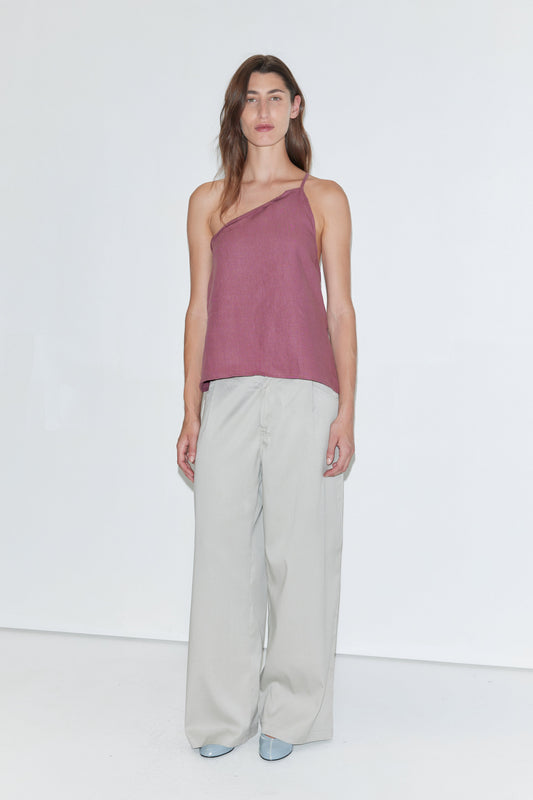 A person stands against a plain white background wearing the Half Halter - Brick linen one-shoulder top from Deiji Studios, paired with loose light gray pants and light blue shoes.