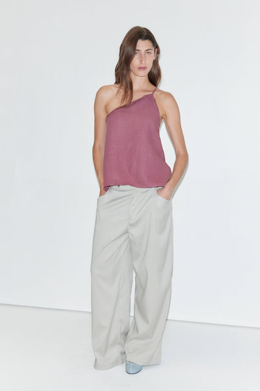 A woman stands against a white background, wearing the Half Halter - Brick linen one-shoulder top from Deiji Studios, wide-legged light gray pants, and blue shoes, with her hands in her pockets.