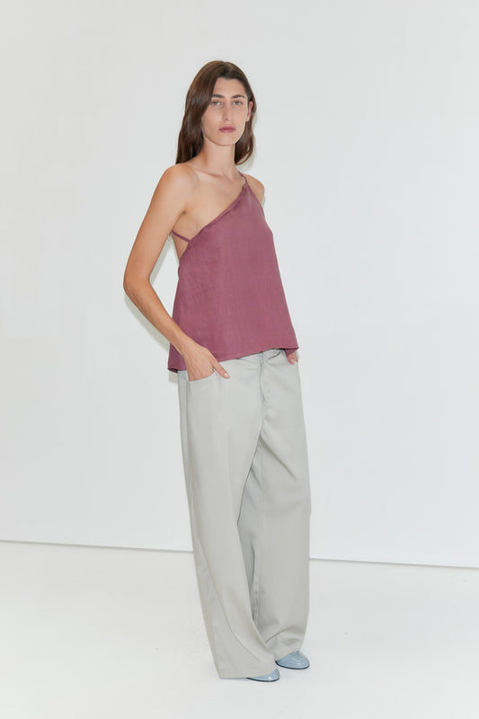 A woman stands against a white background, wearing the "Half Halter - Brick" one-shoulder top by Deiji Studios, loose light gray pants, and light blue shoes. She has long brown hair and her hands in her pockets.