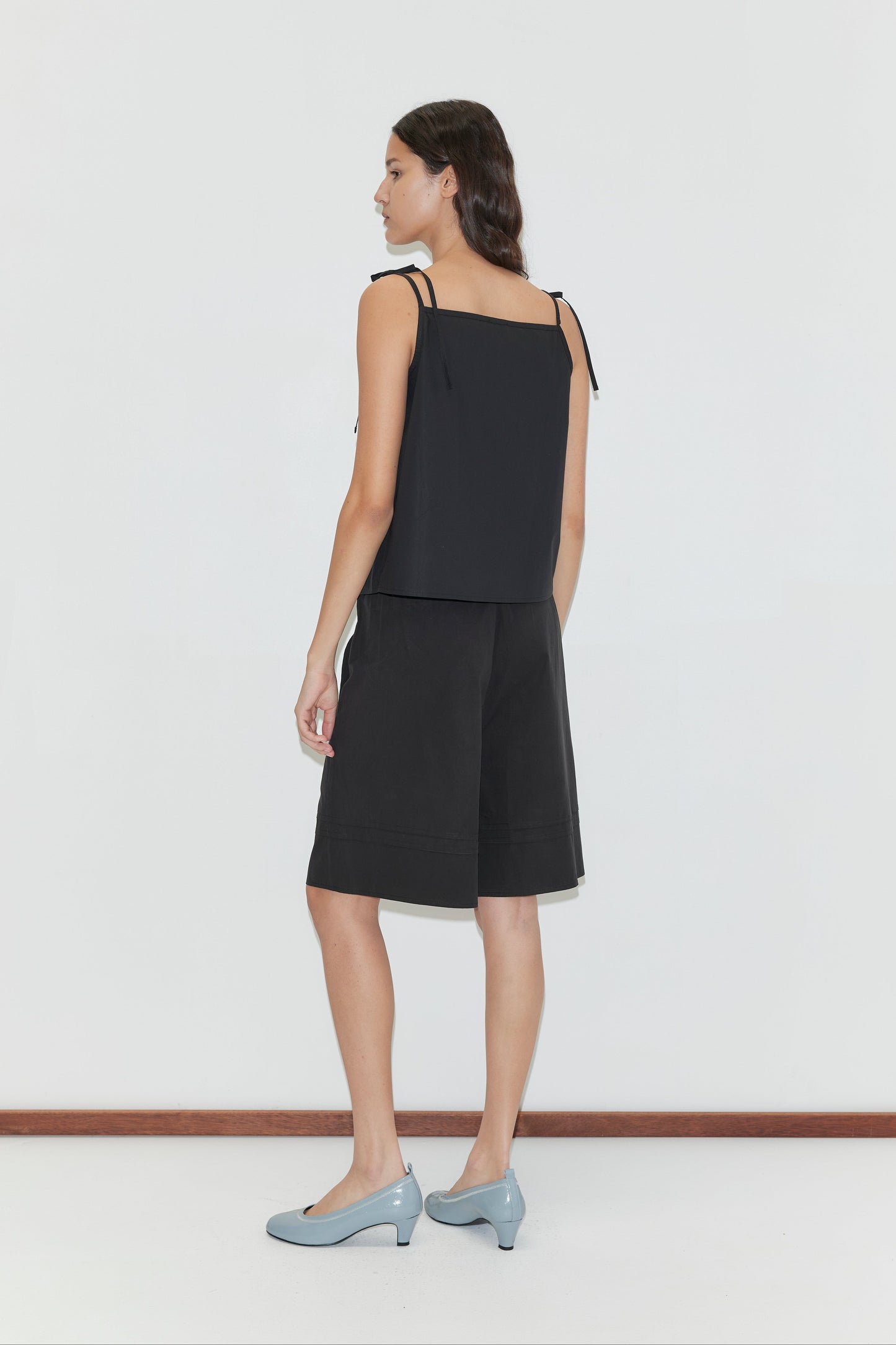 A woman stands against a plain white background wearing the Tied Cami - Black by Deiji Studios, a loose black sleeveless top, paired with black shorts and light blue heels, standing slightly turned away from the camera.