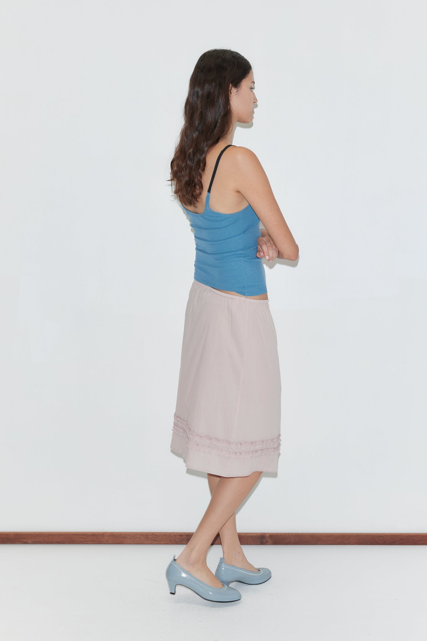 A woman with long brown hair stands in profile against a plain white wall, wearing a blue sleeveless top, the Daytime Skirt - Floss in light pink cotton voile, and blue low-heeled shoes. Her arms are crossed over her chest.
