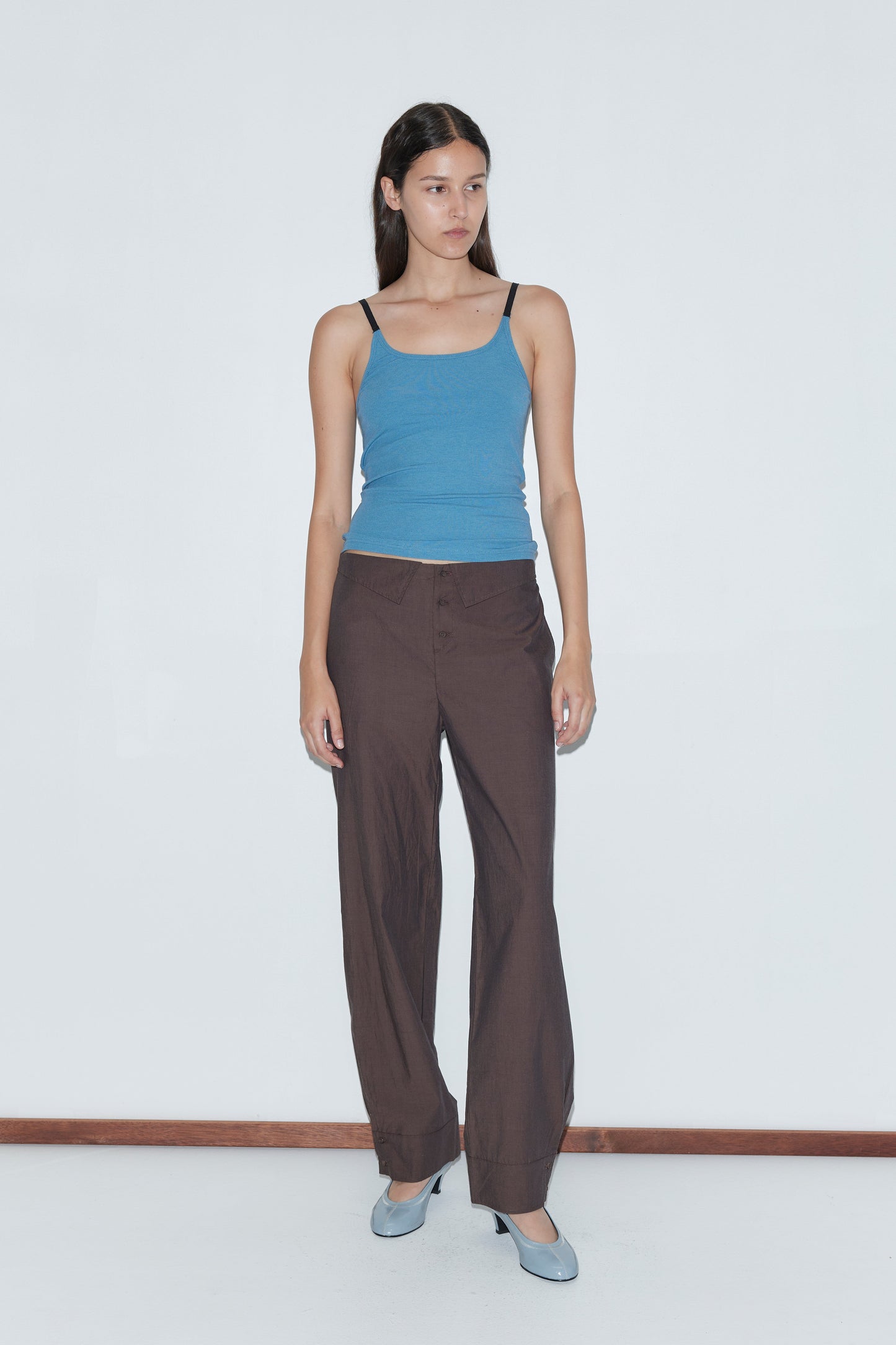 A woman with long dark hair and a neutral expression stands against a white wall, wearing a blue sleeveless top, the Collar Point Pant - Toast, and light blue heels.