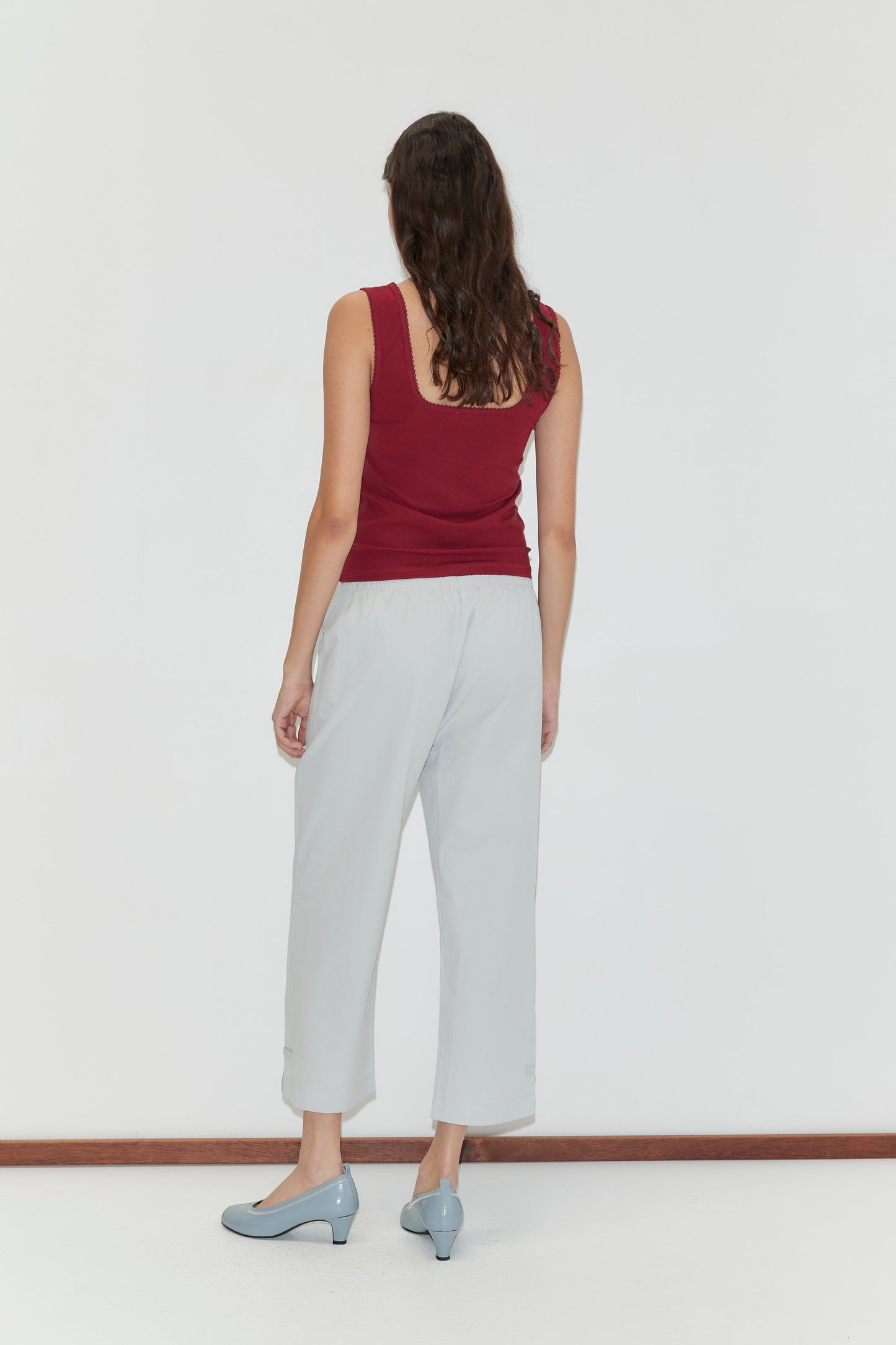 A woman with long brown hair, wearing a sleeveless maroon top and Capri Pant - Baby Blue, stands facing a white wall with her back to the camera, her light blue low-heeled shoes visible.