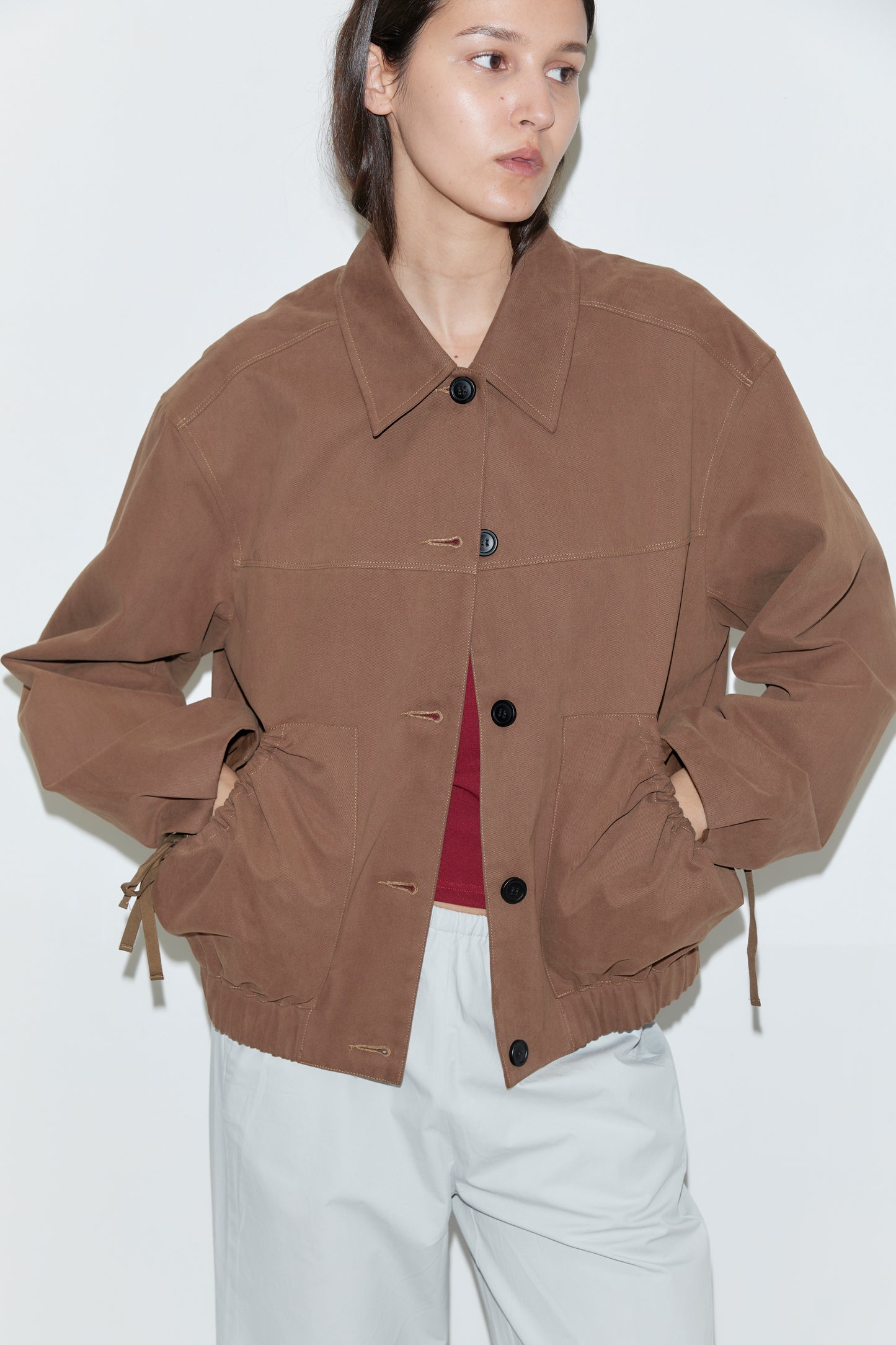A person wears the Button Up Jacket - Brown over a red top and light gray pants, standing against a plain white background with hands in the jacket pockets.