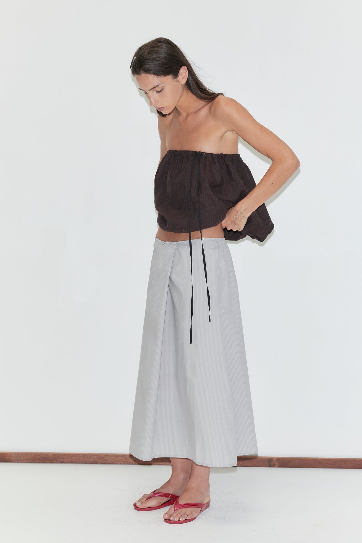 A woman with long dark hair stands in profile against a plain white background, wearing the Bubble Top - Brown Black Stripe by Deiji Studios and a light grey striped wrap skirt with red flip-flops.