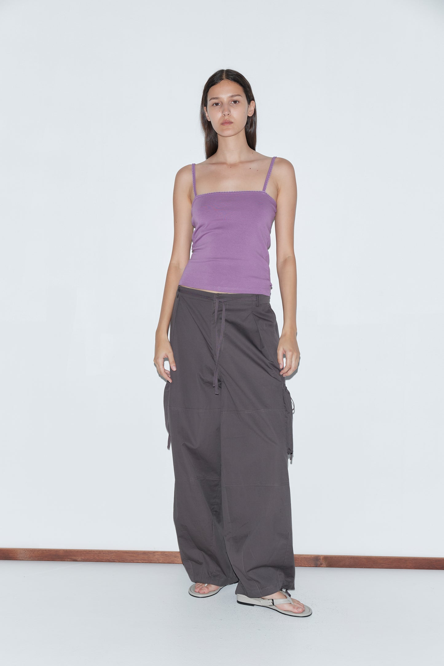 A woman with long, straight hair and a neutral expression stands against a white background, wearing a fitted purple spaghetti strap top, Cargo Pant - Carob by Deiji Studios, and sandals.