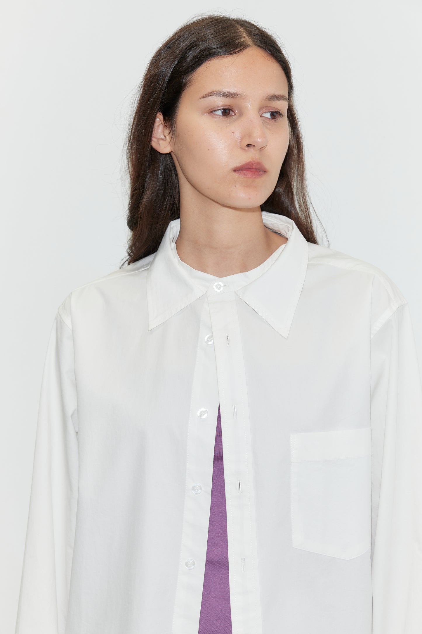 A woman with long brown hair wears the Stand Shirt - White from Deiji Studios over a purple top, standing against a plain white background and gazing slightly to the side.
