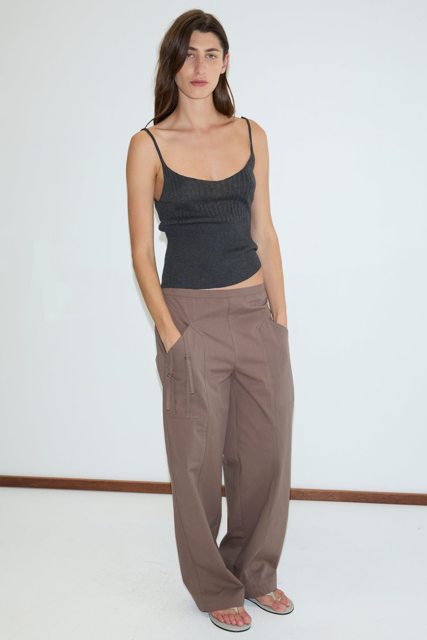 A woman with long brown hair and a neutral expression stands against a plain white wall wearing Deiji Studios’ dark gray sleeveless ribbed top, Adjust Pocket Pant in Sawdust, and sandals. She has her hands in the pockets of the pants.