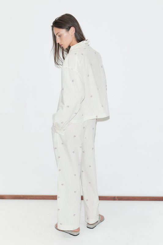 A person stands sideways against a white background, wearing the Ease Trouser - Floral Stripe pajama set, which features a loose fit with straight-leg trousers adorned with small, subtle floral stripe patterns. Their arms hang relaxed by their sides, and their long brown hair falls forward as they look down slightly.