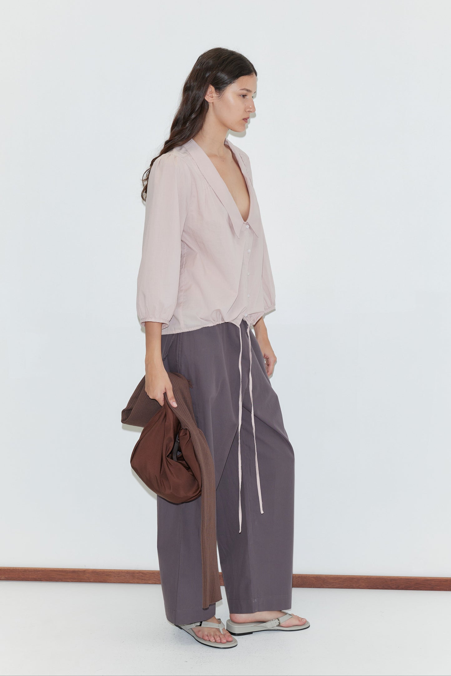 A woman with long dark hair wears a loose pale pink blouse and wide-leg gray drawstring pants from Deiji Studios, holding the Knit Strap Bag - Hazel and a jacket, in sandals, standing against a plain white background.