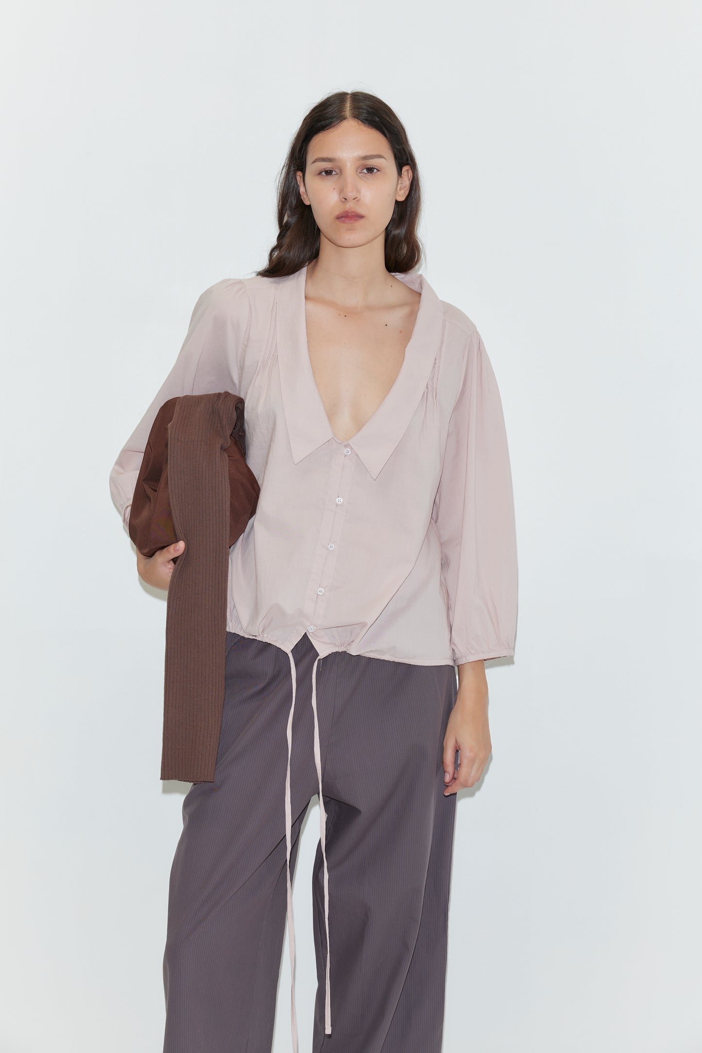 A woman with long dark hair wears a loose, light mauve blouse and dark pants, holding a brown garment over her arm. She accessorizes with the Knit Strap Bag - Hazel and stands against a plain white background.