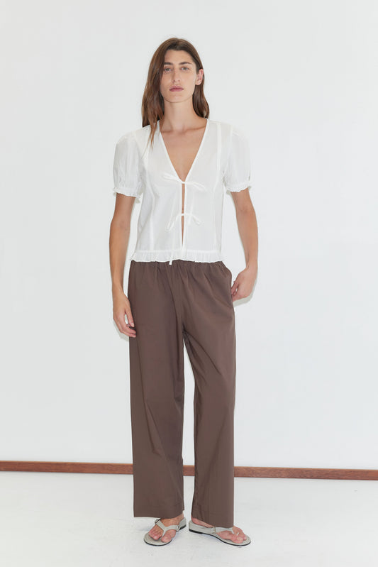 A person stands against a plain white background wearing a white short-sleeve V-neck blouse with front ties, Ease Trouser - Chocolate by Deiji Studios, and beige flip-flops.