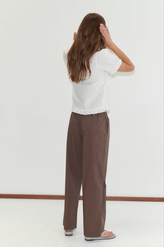 A person with long brown hair, wearing a white short-sleeve blouse, the Deiji Studios Ease Trouser in Chocolate, and sandals stands facing a white wall with hands raised to their head.