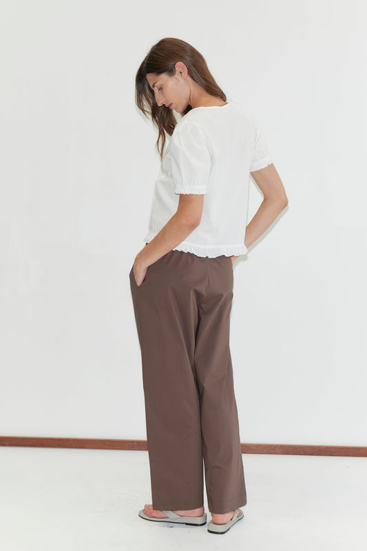 A woman stands sideways against a white background, looking down. She wears a white short-sleeved blouse, Deiji Studios Ease Trouser - Chocolate, and gray sandals, with one hand in her pocket.