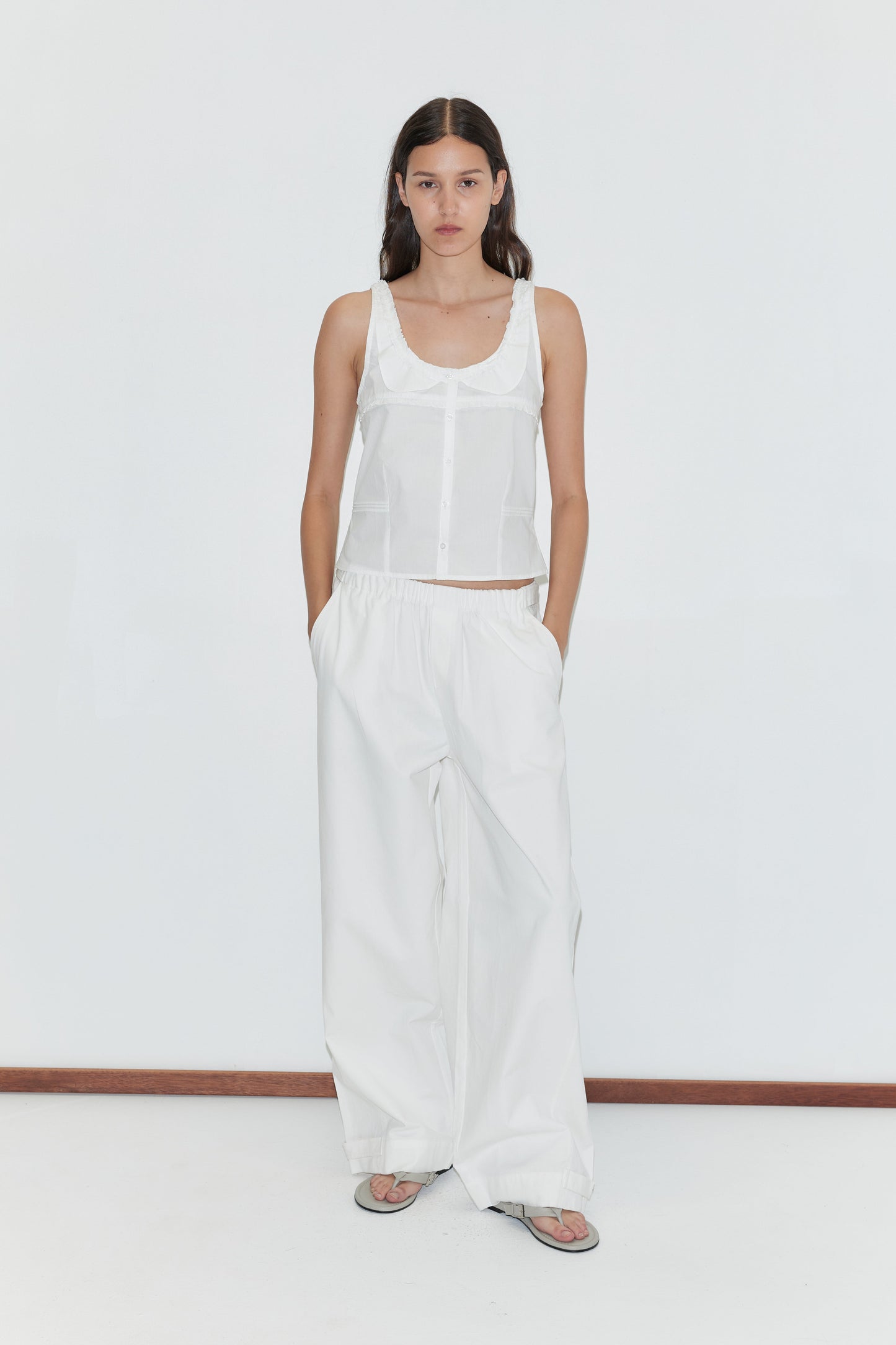 A woman with long brown hair stands against a white background wearing a sleeveless white button-up top, gray sandals, and the Snap Tab Pant - White by Deiji Studios, with her hands tucked casually in her pockets.