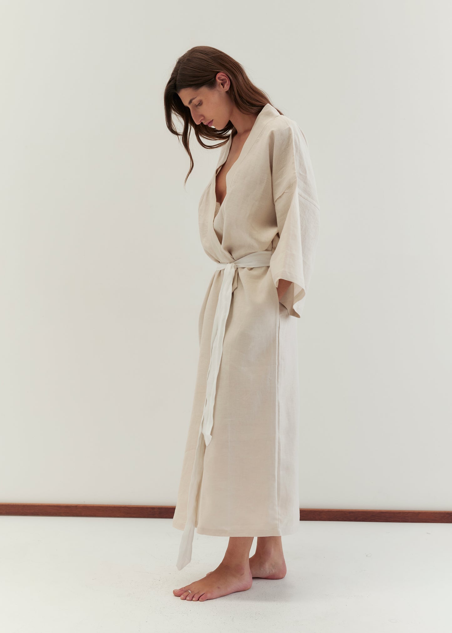 A barefoot woman with long brown hair stands against a plain white background, wearing the Deiji Studios 02 robe in oatmeal, loosely tied at the waist with a linen belt. She looks down with her arms relaxed by her sides.