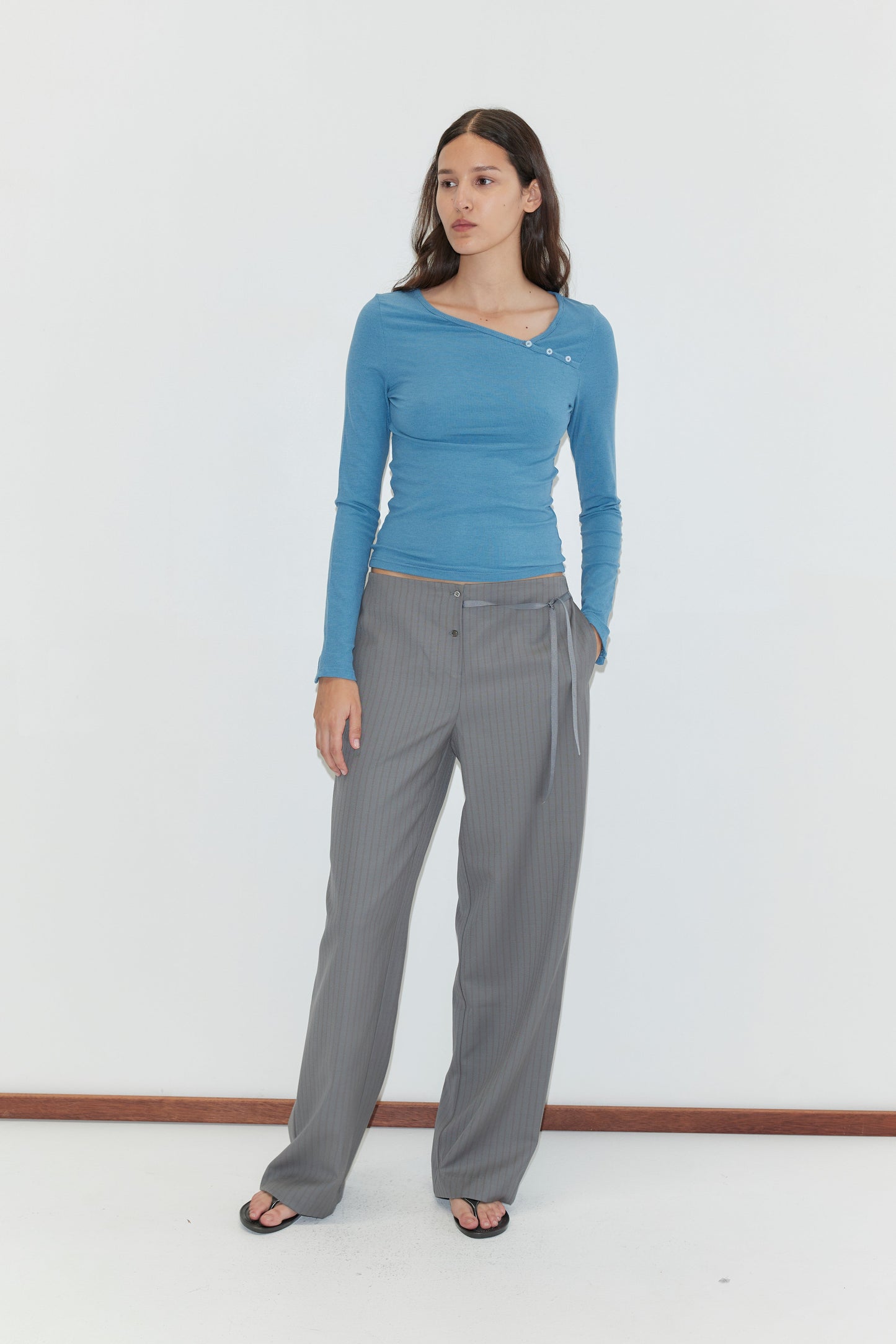 A woman with long brown hair and a neutral expression stands against a white background wearing a fitted blue long-sleeve top, black sandals, and the Boxer Pant - Grey Stripe: wool wide-leg pinstriped trousers with an elasticated waistband and belt.