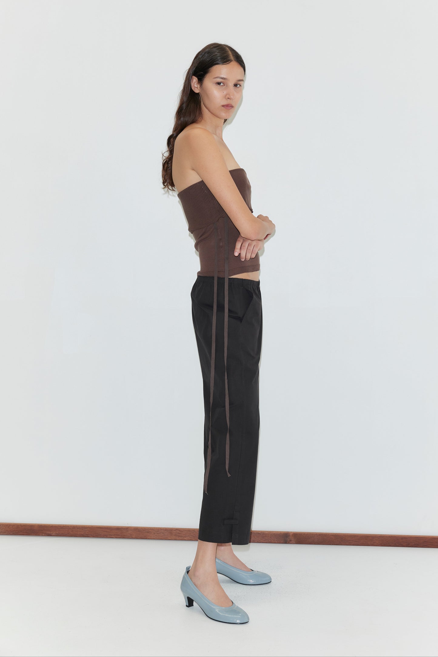 A woman with long dark hair stands sideways against a white wall, arms crossed, wearing a brown strapless top, Capri Pant - Black from Deiji Studios, and light blue heeled shoes.