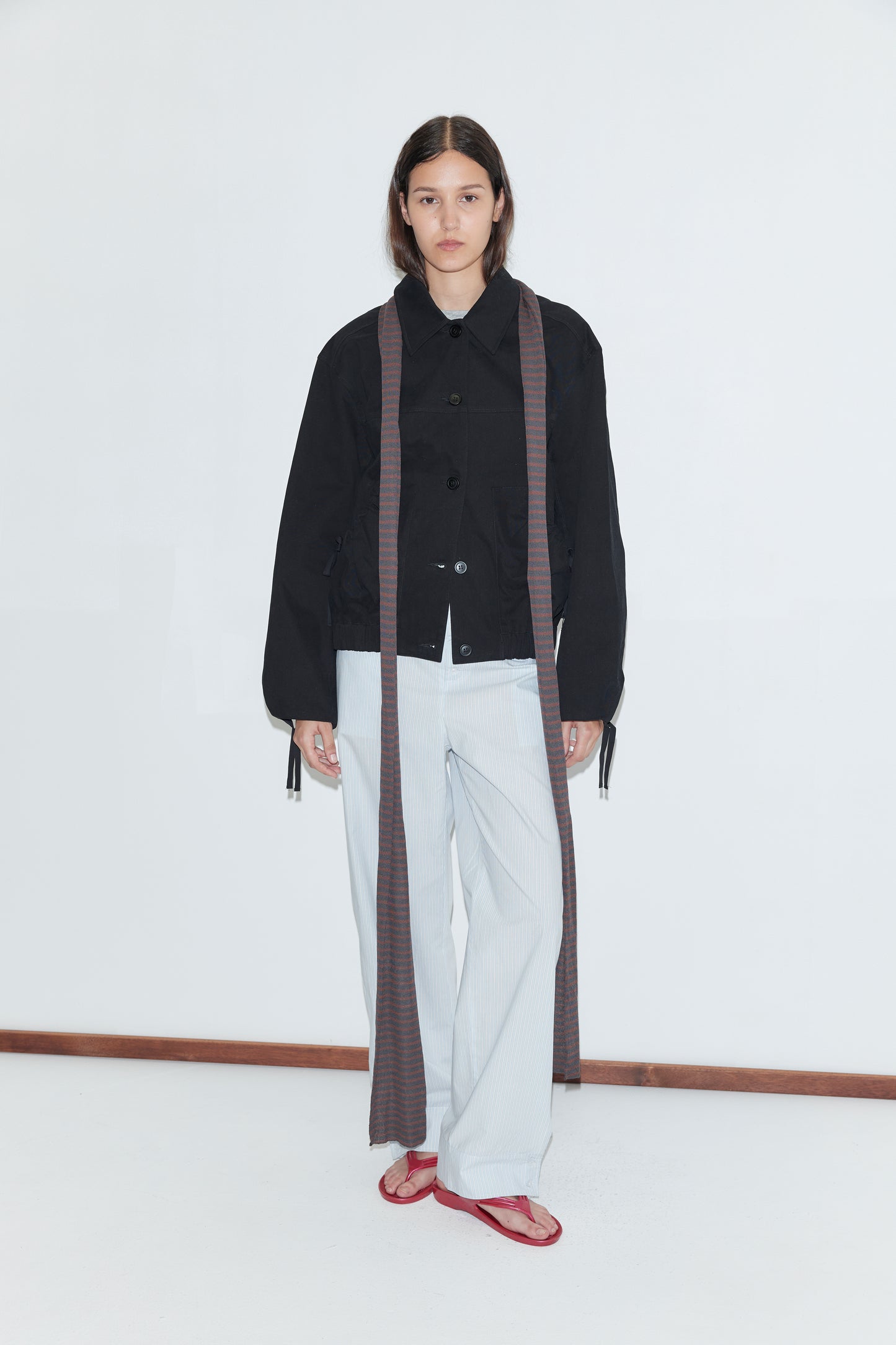 A person with long dark hair and a neutral expression stands against a white background wearing the Button Up Jacket - Black by Deiji Studios, light striped pants, a long scarf draped on both sides, and red sandals.