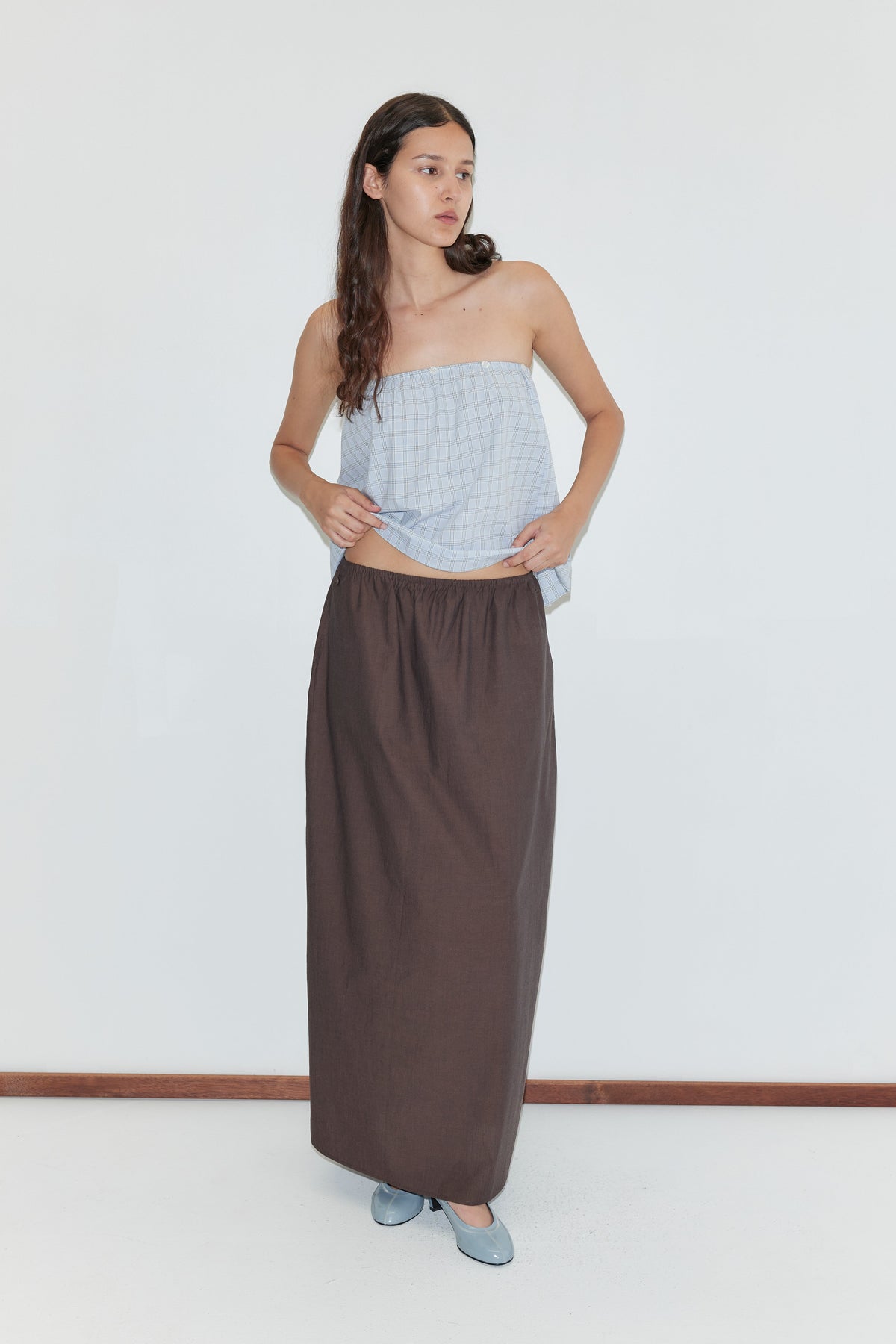 A woman with long, wavy hair stands indoors against a plain white background wearing the Way Skirt in Toast, a strapless light blue plaid top, and blue flat shoes. She looks to the side.