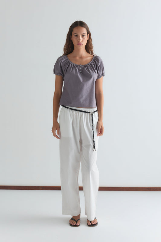 A woman with long, wavy brown hair and a neutral expression stands against a white background wearing the Poplin T Shirt - Currant by Deiji Studios, paired with loose white pants, a black belt, and black sandals.