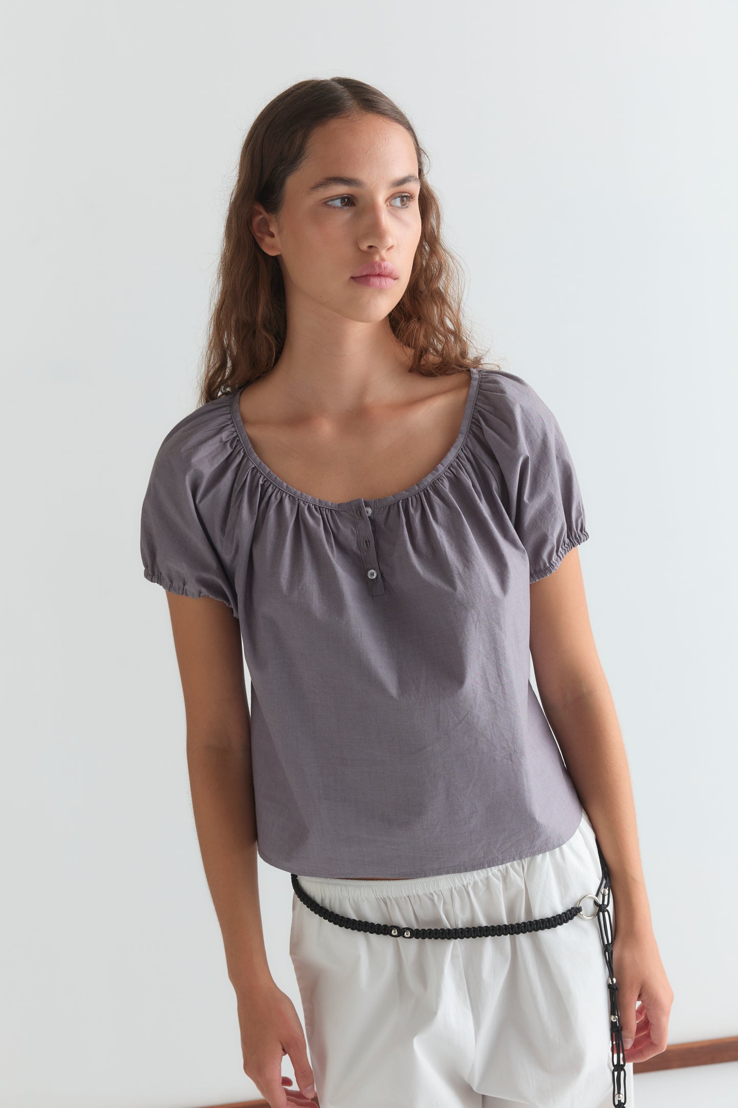 A woman with long brown hair wears the Deiji Studios Poplin T Shirt - Currant in a cropped relaxed fit and white shorts with a black braided belt, standing against a plain white background and looking to the side.