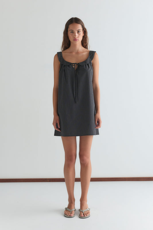 A woman stands against a white wall, wearing the Rounded Neck Dress - Mono stripe by Deiji Studios—a sleeveless, navy and white striped mini dress with front tie detail—paired with silver sandals. She faces forward with a neutral expression.