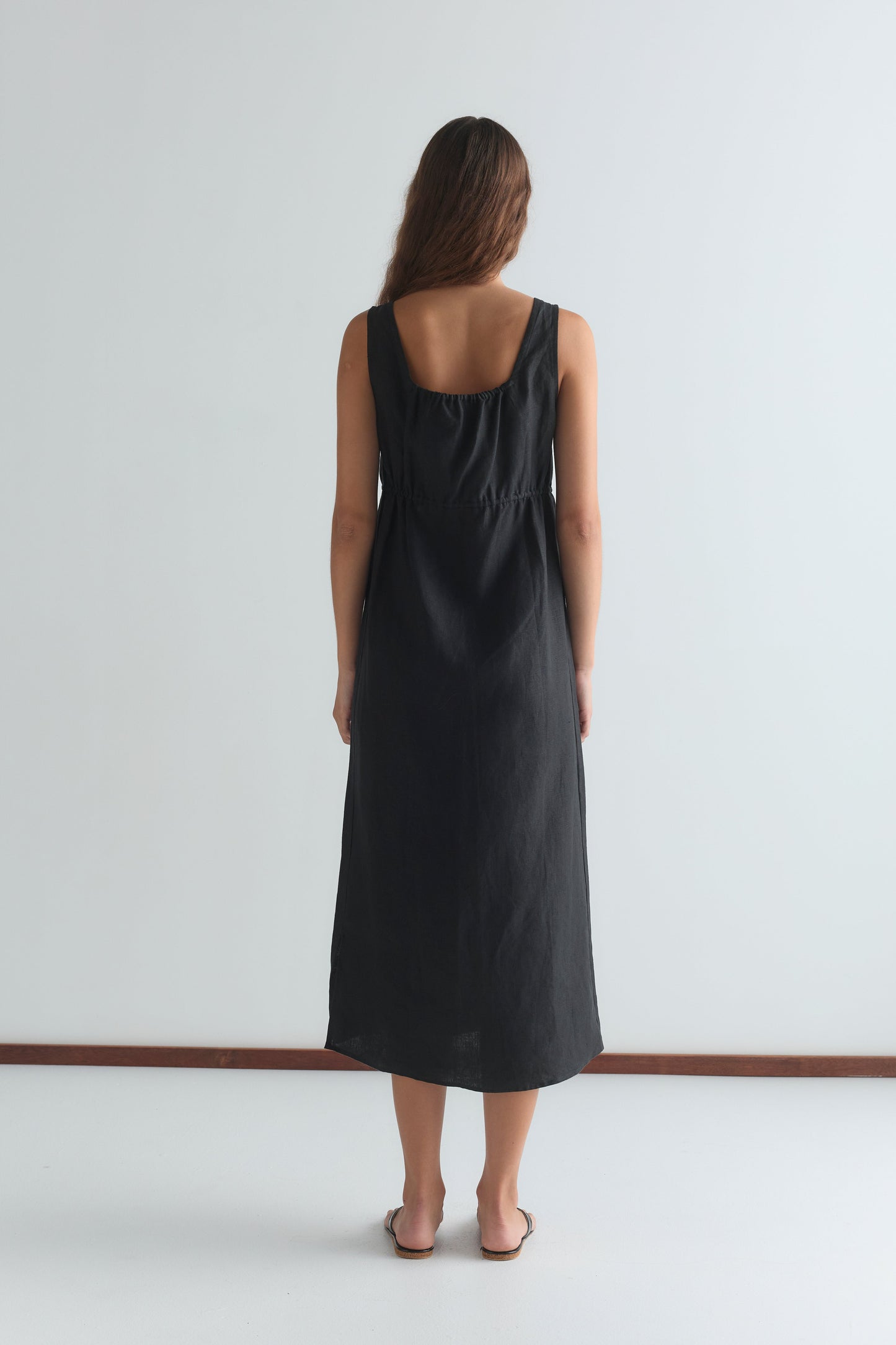 A woman with long brown hair stands barefoot, facing a white wall and wearing the Sleeveless Cord Dress - Black, which falls to her ankles in an effortlessly relaxed midi style.