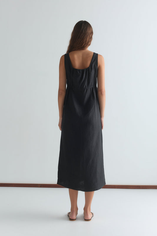 A woman with long brown hair stands barefoot, facing a white wall and wearing the Sleeveless Cord Dress - Black, which falls to her ankles in an effortlessly relaxed midi style.