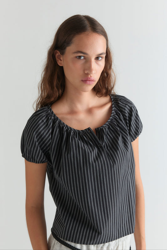 A young woman with long brown hair wears the Poplin T Shirt - Mono Stripe by Deiji Studios and light-colored pants, standing against a plain light background.