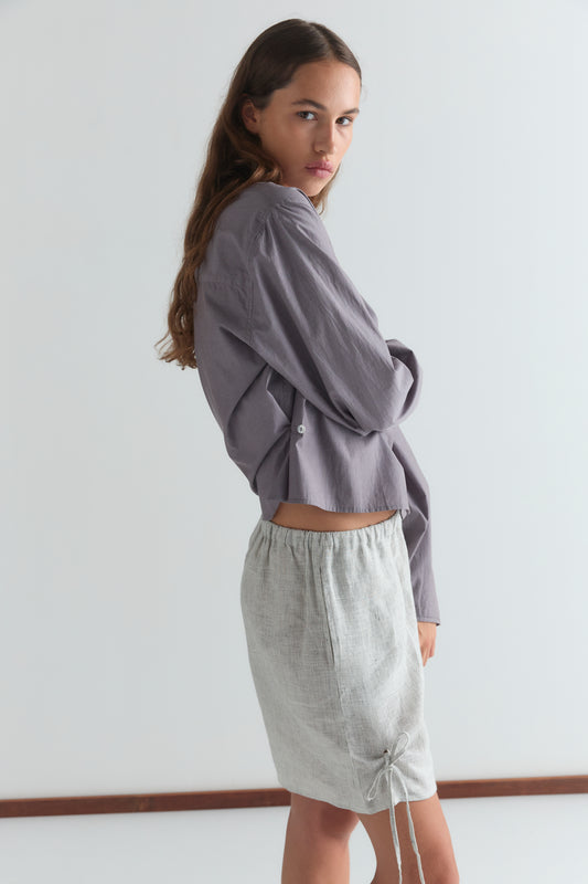 A woman with long brown hair wears a loose, long-sleeved lavender shirt and Fine Stripe Eyelet Shorts, standing sideways and looking over her shoulder against a plain white background.