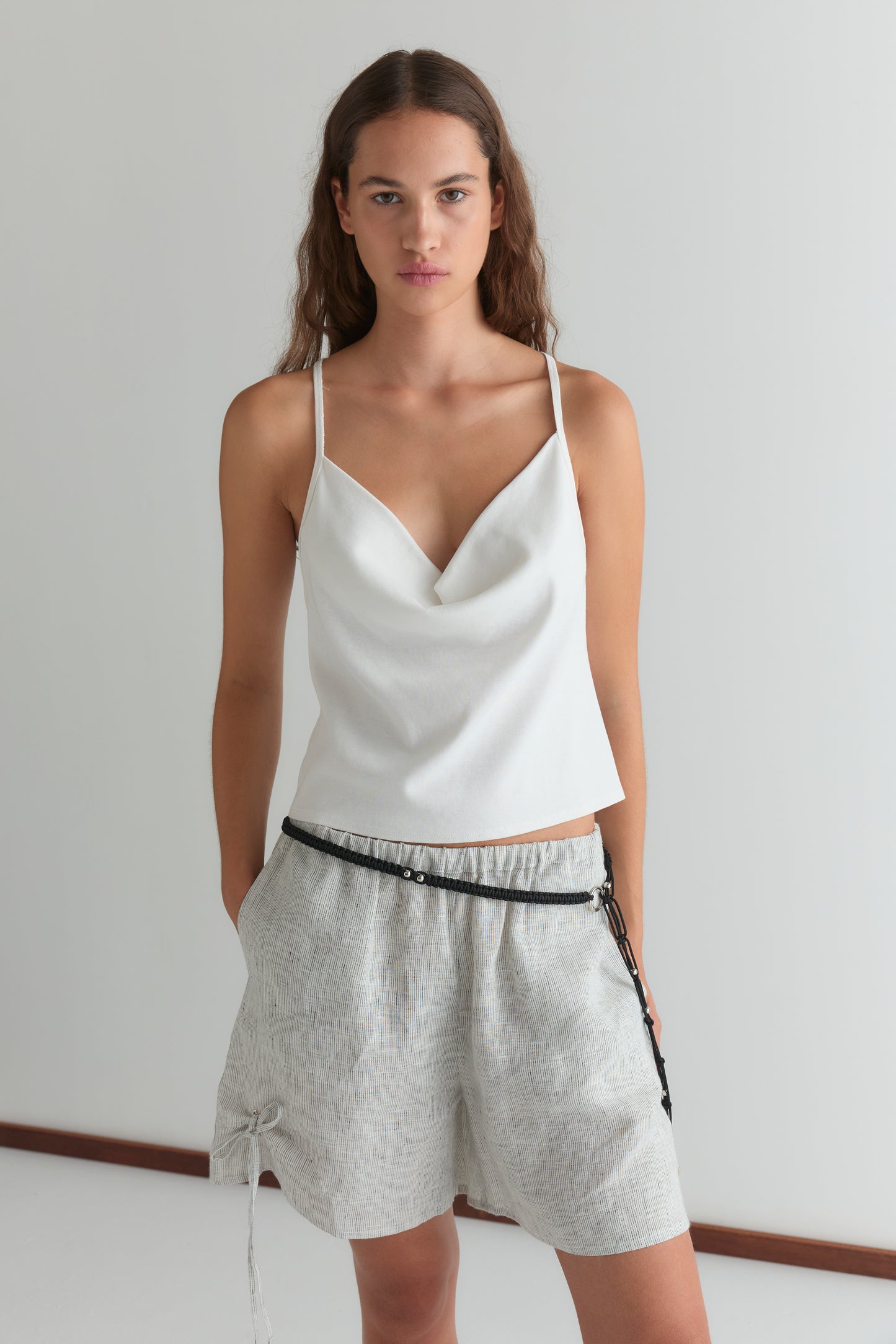 A woman with long brown hair stands against a white wall, wearing the Drape Jersey Top in white and loose light gray shorts with a black belt. Her hands are in her pockets as she gazes directly at the camera.