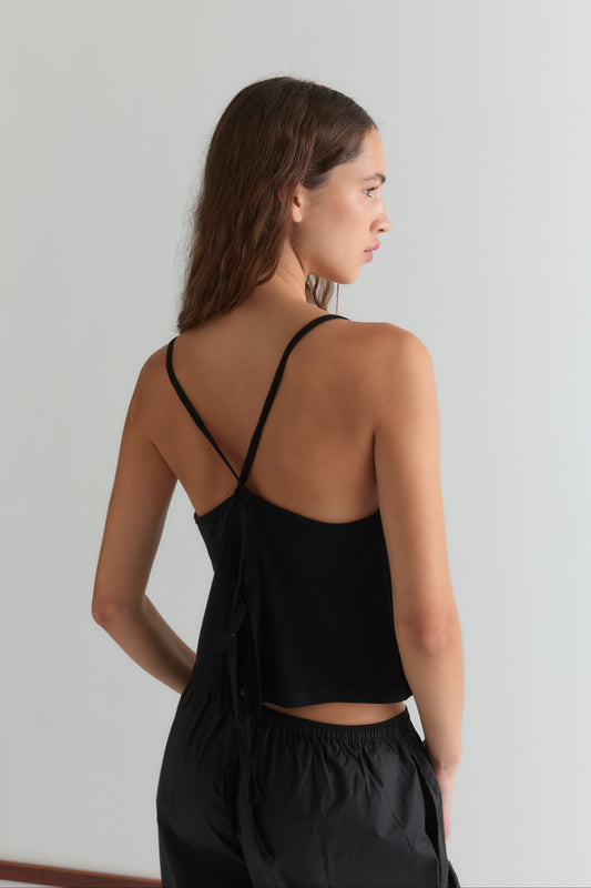 A woman with wavy brown hair stands sideways, facing away, wearing a black Drape Jersey Top by Deiji Studios with an open back and black pants against a plain light background.