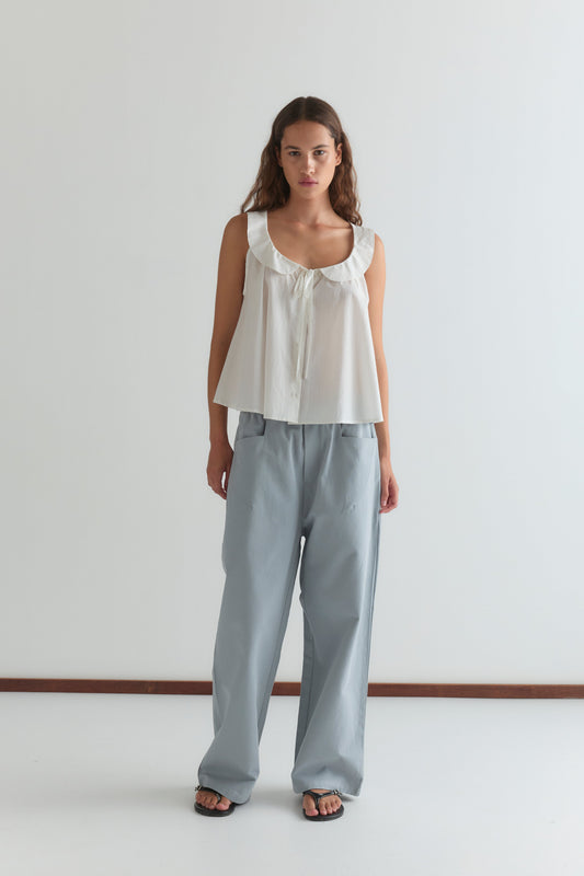 A woman with long brown hair stands against a white wall, wearing the Round Collar Top - White Texture by Deiji Studios, styled with light blue loose-fitting pants and black sandals.