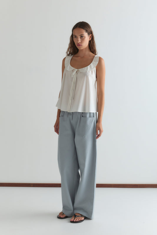 A woman with long brown hair and a neutral expression stands against a white wall wearing the Round Collar Top - White Texture, featuring a ruffled collar and tie detail, paired with light blue wide-leg pants and black sandals.