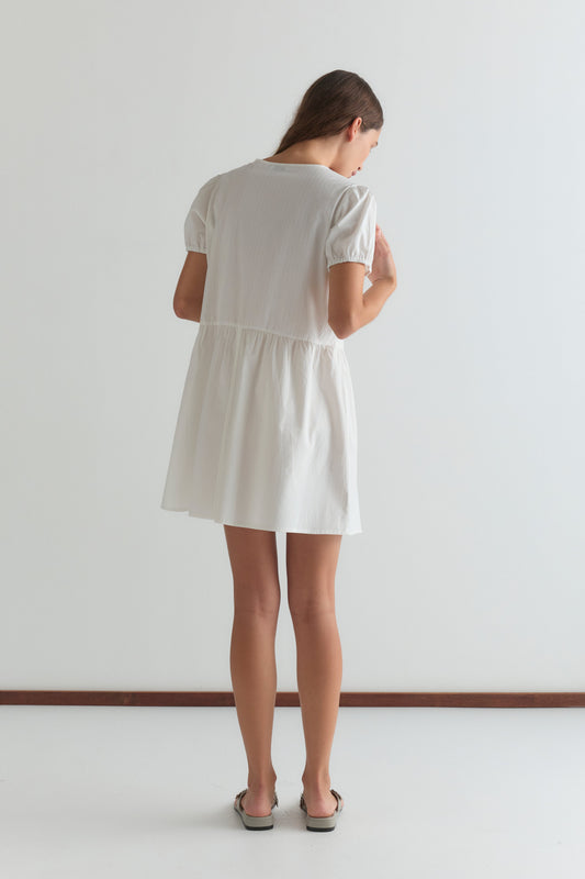 A woman with long brown hair faces away, wearing the V Pleat Dress - White Texture from Deiji Studios with short sleeves and flat sandals, set against a plain white background.