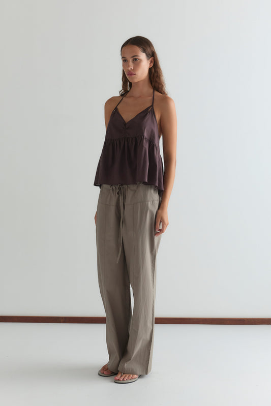 A woman with long brown hair wears the Keyhole Halter Top - Wine by Deiji Studios, featuring adjustable straps, paired with loose beige pants and flip-flops, standing against a plain white wall.