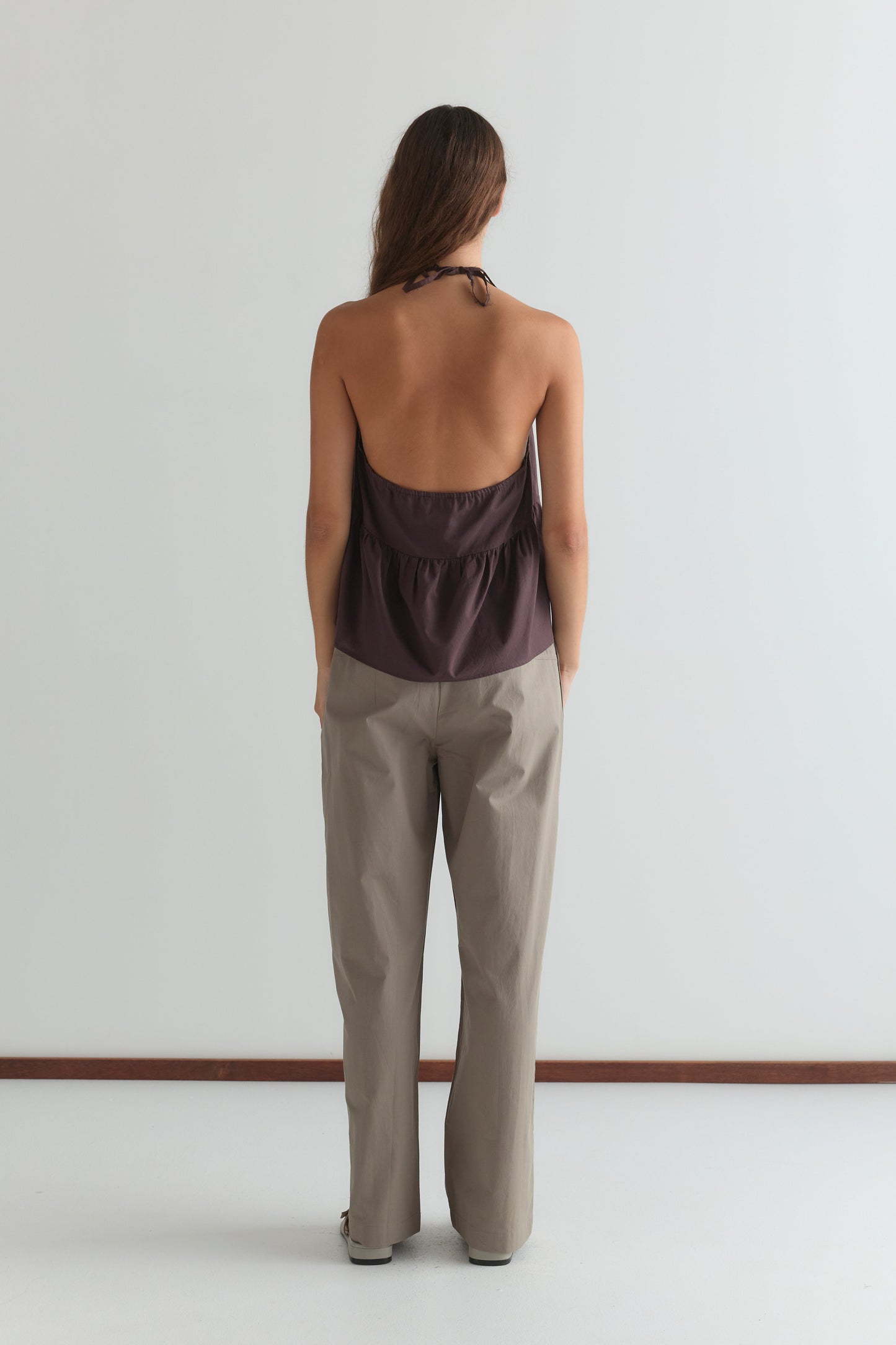 A person with long brown hair stands facing away, wearing the Keyhole Halter Top - Wine by Deiji Studios with adjustable straps and light-colored loose pants in a minimalist space with white walls and floor.