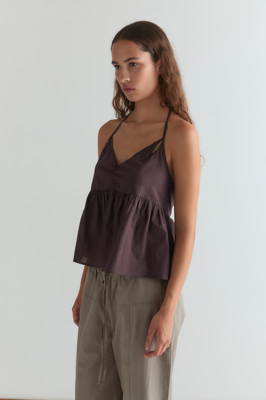 A young woman with long brown hair wears the Keyhole Halter Top - Wine by Deiji Studios, featuring adjustable straps, paired with loose beige pants, standing against a plain light background.