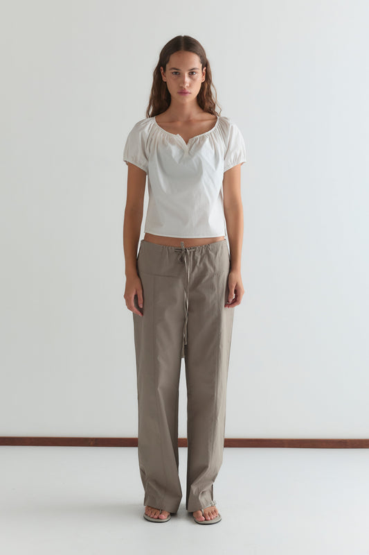 A woman stands facing forward against a plain white background, wearing the Poplin T Shirt - White by Deiji Studios, paired with loose beige trousers and sandals.