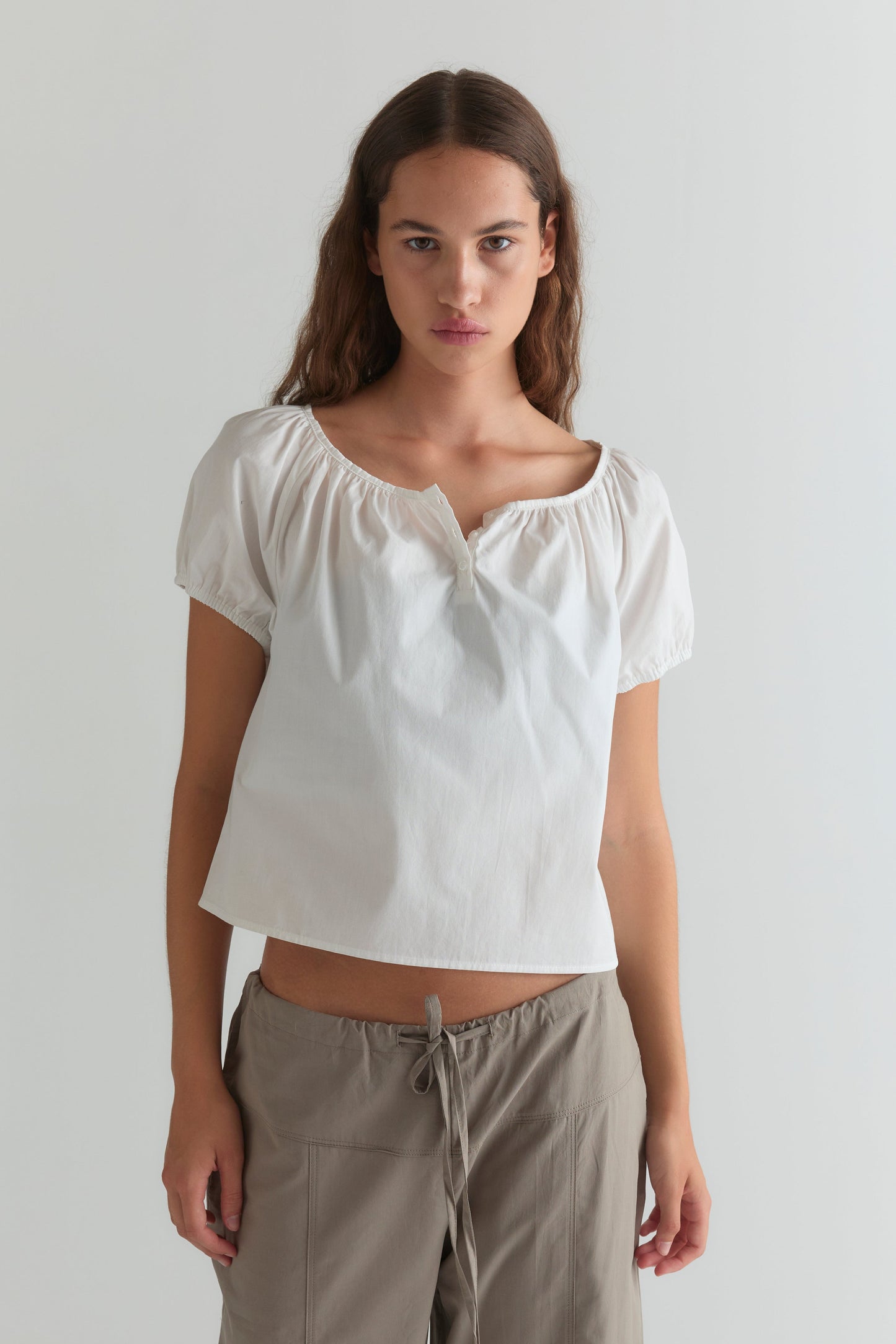 A person with long brown hair wears the Deiji Studios Poplin T Shirt - White and beige drawstring pants, standing against a plain light-colored wall.
