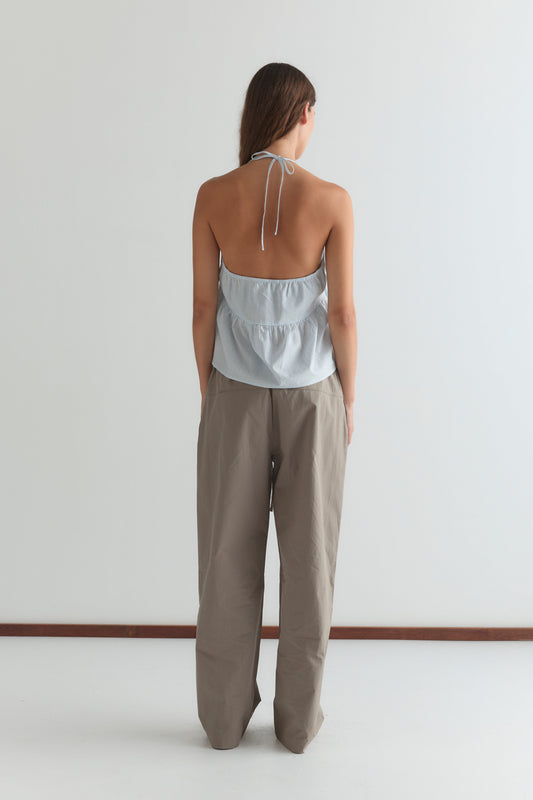 A woman stands facing away, wearing a light blue halter-top and loose, high-waisted Seam Pant - Light Khaki from Deiji Studios, against a plain white wall.