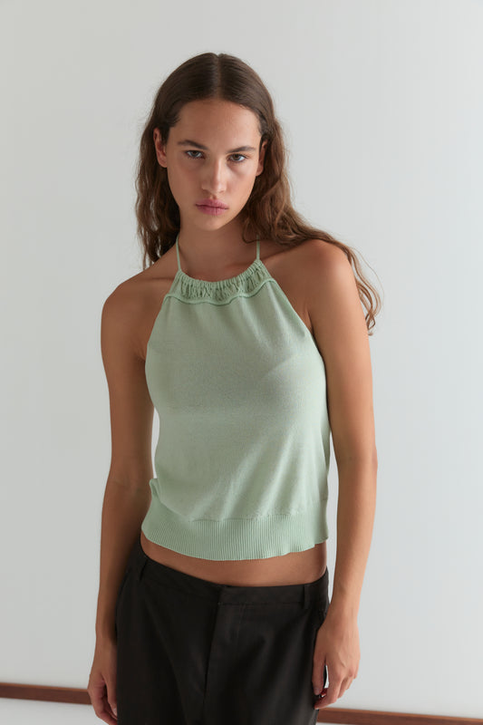 A young woman with long brown hair wears the Deiji Studios Drawcord Knit Top in Mint, featuring an adjustable ruched neckline, paired with dark pants. She stands against a white background, looking at the camera with a neutral expression.