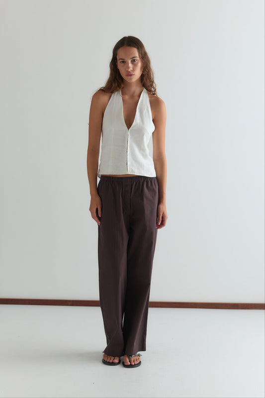 A woman with long hair stands against a white wall, wearing Deiji Studios’ Close Halter Top in white—a sleeveless halter style with a deep V neckline—paired with loose dark brown wide-leg pants. She gazes ahead with a neutral expression.