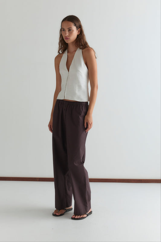 A woman stands against a plain white wall, wearing the Close Halter Top - White from Deiji Studios, loose dark brown pants, and black sandals. She has long, wavy hair and a neutral expression.