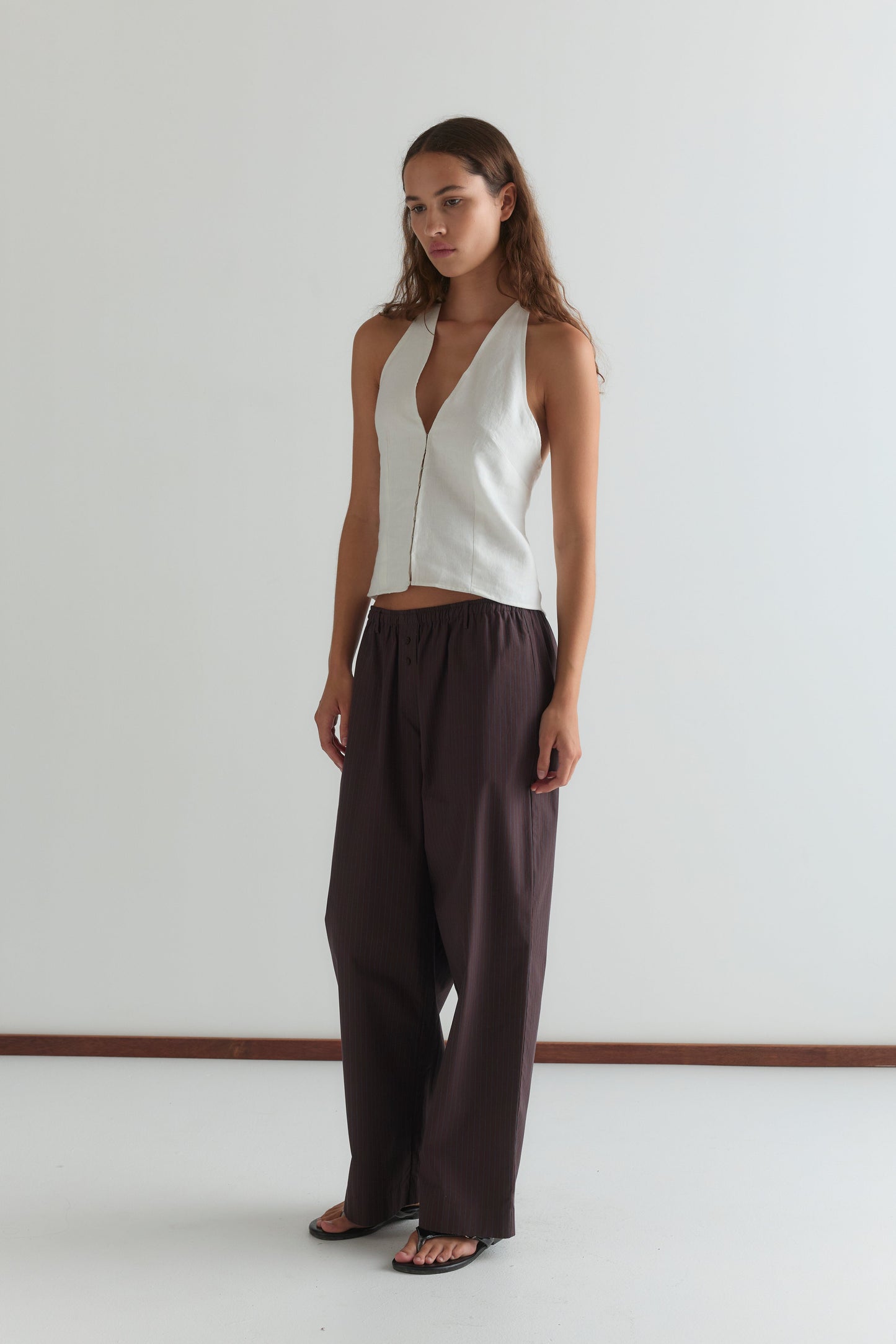 A woman with long brown hair and a neutral expression stands in a plain room wearing a sleeveless white V-neck top, black sandals, and Deiji Studios’ Double Button Pant - Brown Stripe.